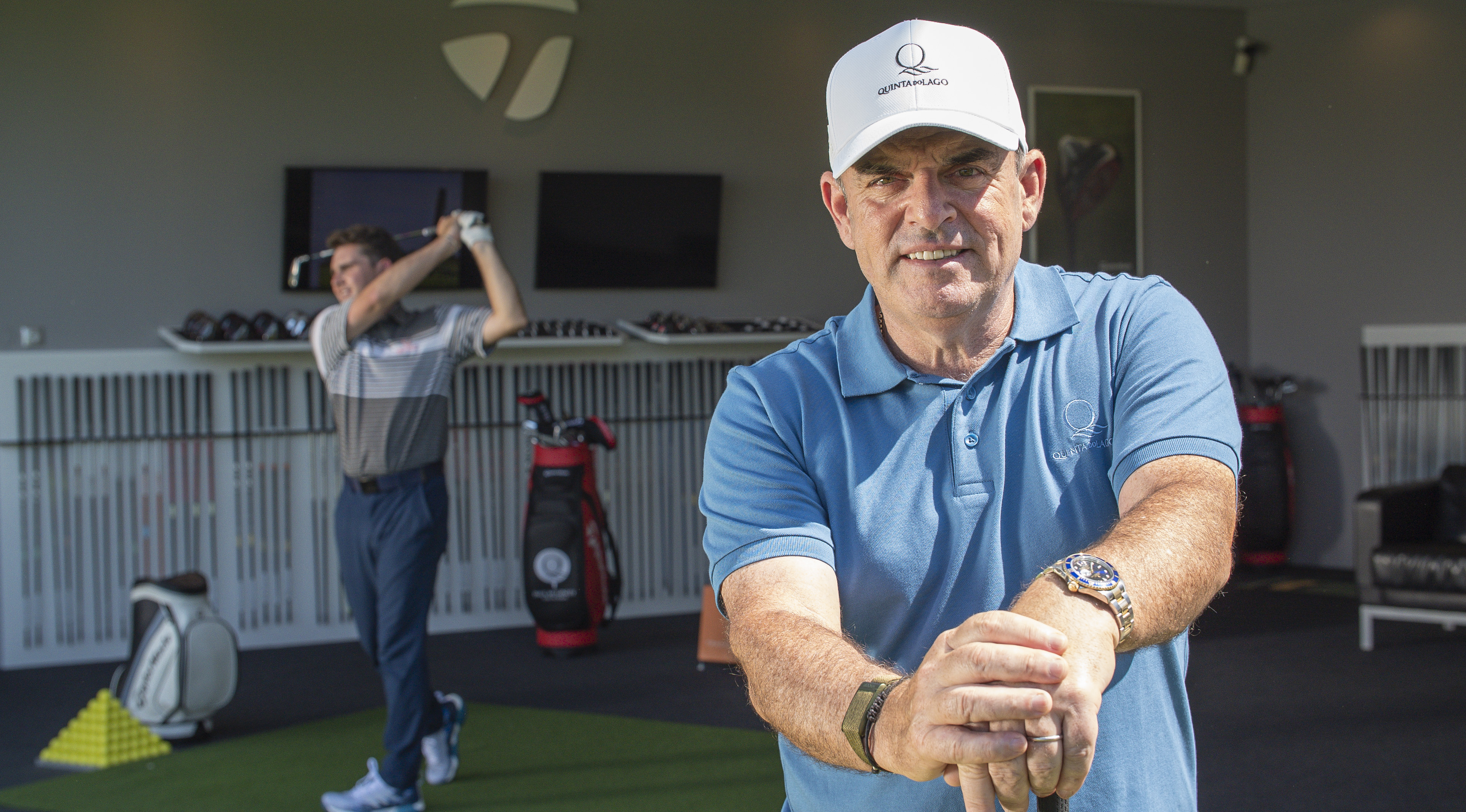 Golf lesson at the Paul McGinley Golf Academy in Quinta do Lago, with a professional instructor in a blue shirt demonstrating swing technique, while a student practices in the background. Premium golf coaching in the Algarve, Portugal