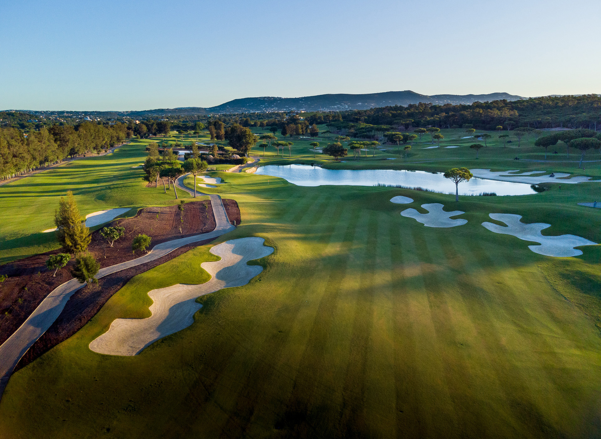 Aerial view of Laranjal Golf Course at Quinta do Lago, Portugal, featuring immaculate greens, strategic sand bunkers, and a scenic pond framed by orange groves and pine trees under clear blue skies. Premium golf experience in the Algarve.